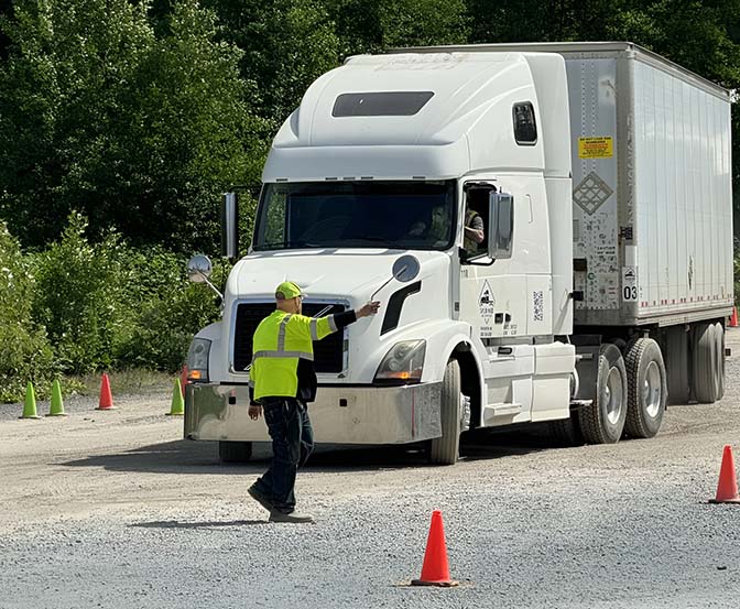 DDriver instruction - Taylor Made Truck Driving School A person wearing a bright, easy-to-see vest and hat is waving to a big white truck. The truck is driving on a rocky road with orange cones all around, and there are trees and bushes in the background. - Taylor Made Truck Driving School