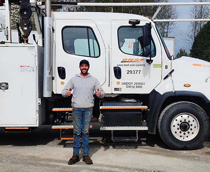 Job placement support - Taylor Made Truck Driving School A person wearing a gray hoodie and jeans is standing and smiling in front of a BNSF work truck. They are giving a thumbs-up with both hands. You can see the truck's logo and some numbers on its side. - Taylor Made Truck Driving School