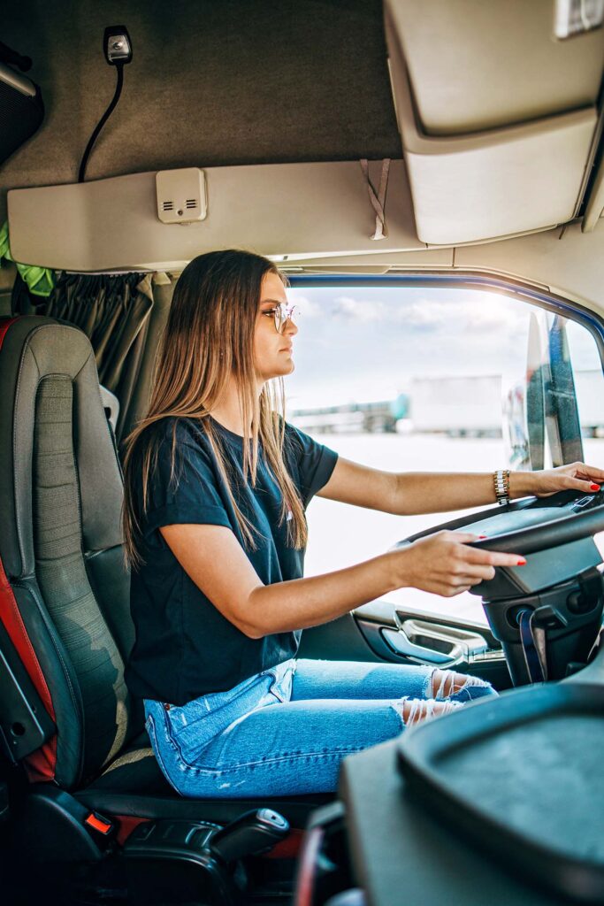 A person with long hair and glasses is sitting in the driver’s seat of a truck. They are holding the steering wheel and looking straight ahead. - Taylor Made Truck Driving School A person with long hair and glasses is sitting in the driver’s seat of a truck. They are holding the steering wheel and looking straight ahead. - Taylor Made Truck Driving School