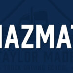 Imagine a big, dark sign. On this sign, there's a very important word written in **BIG WHITE LETTERS** that say "HAZMAT." Underneath these big letters, you can just barely see some smaller words that say "Taylor Made Truck Driving School. - Taylor Made Truck Driving School