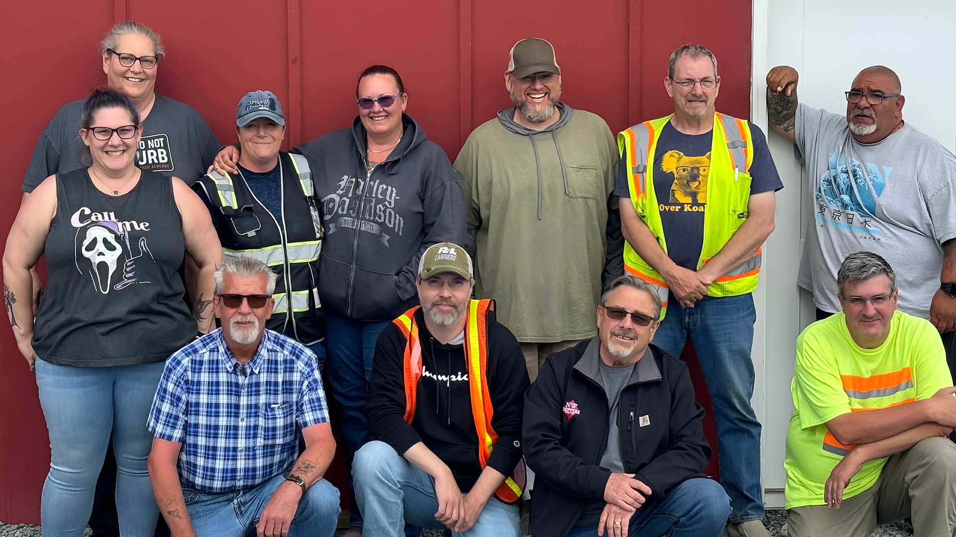OOur instructors - Taylor Made Truck Driving School Truck driving school instructors. They are in front of a red and white wall. Some people are standing up, while others are squatting or sitting on the ground. - Taylor Made Truck Driving School
